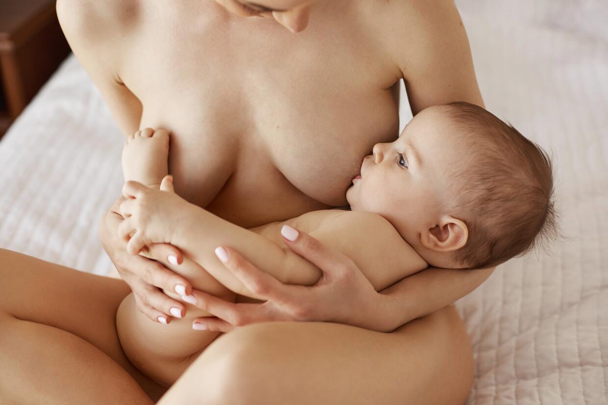 Stock photo 657764986 - Young beautiful naked mom breastfeeding hugging her  newborn baby smiling sitting on bed at home.